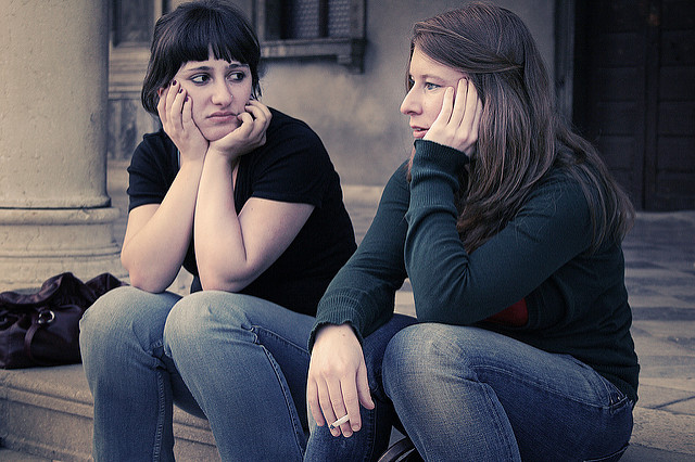 Two women sitting outside of a building, smoking a cigarette