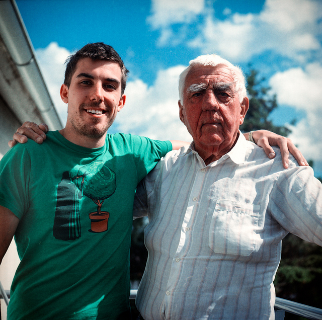 A young adult standing with his arm around his grandpa