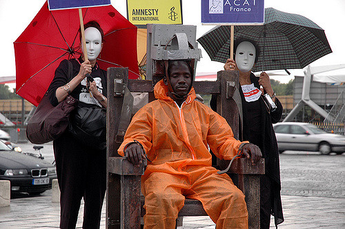 Three people protesting the death penalty and how it is racially discriminative. A black man sits in a mock electric chair, while two women wearing white masks stand watching.
