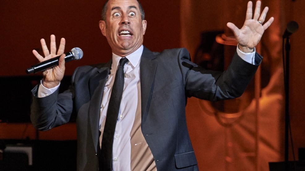 Jerry Seinfeld looking taken aback, big eyes, open hands, mic under his thumb